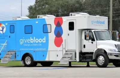 A large white and blue mobile blood donation truck is parked on a paved area, with giveblood and Change a Childs Life written on the side. Three entry doors with steps lead into the vehicle. Children's Hospital Bloodmobile