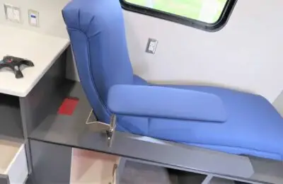 A blue medical recliner chair with an armrest positioned next to a desk and power outlets, inside a bright room with a window.