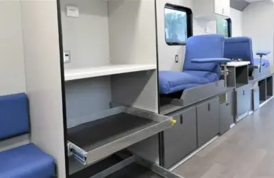 Interior of a mobile medical unit with blue chairs, fold-out metal trays, storage cabinets, and a window along the wall. The environment appears clean and organized with gray flooring.
