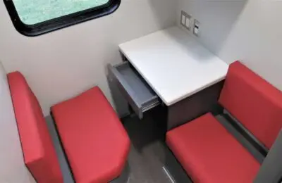 A small dining or work area with a white square table, two red cushioned bench seats, a window, and an open drawer under the table, inside a compact room.