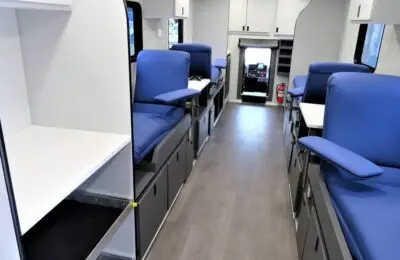 Interior of a mobile medical unit with blue cushioned seats on both sides, white cabinets, and work surfaces. A fire extinguisher is visible near the front drivers area. The floor is gray and the space is well-lit.