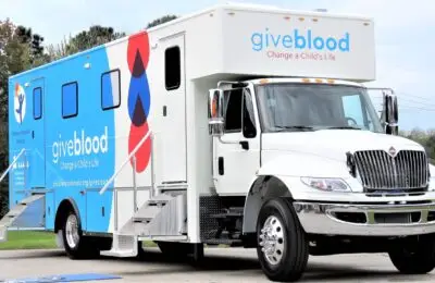 A white and blue blood donation mobile unit with the words giveblood Change a Child’s Life on the side is parked outdoors. Two sets of stairs provide access to the vehicle’s doors. Children's Hospital Bloodmobile