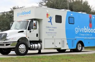 A white and blue mobile blood donor truck with give blood and Childrens Hospital Colorado written on the side, parked on pavement with trees and power lines in the background. Children's Hospital Bloodmobile