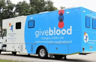 A large blue and white blood donation mobile unit is parked outdoors. The side reads “give blood. Change a Child’s Life” along with the Children’s Hospital Colorado logo and website. Children's Hospital Bloodmobile