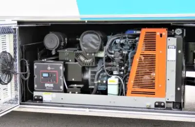 A close-up view of a generator compartment in a vehicle, showing the generator unit, control panel, vents, wiring, and other electrical components. The compartment is open, exposing the machinery inside. Children's Hospital Bloodmobile