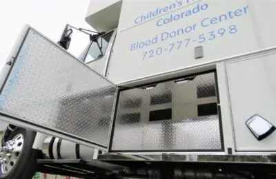 A close-up of an open storage compartment on a white blood donor center vehicle, with diamond plate doors, partial text, and a phone number visible on the side. Children's Hospital Bloodmobile