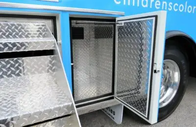 A close-up view of a blue vehicle with metal diamond-plate storage compartments, one with its door open and another with a pull-out stair step extended. Part of a wheel and partial text are visible on the vehicle’s side. Children's Hospital Bloodmobile