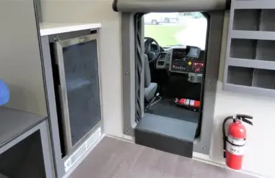 View from inside a vehicle showing the driver’s seat, dashboard controls, a fire extinguisher on the floor and wall, a refrigerator, shelving, and an open doorway leading outside.