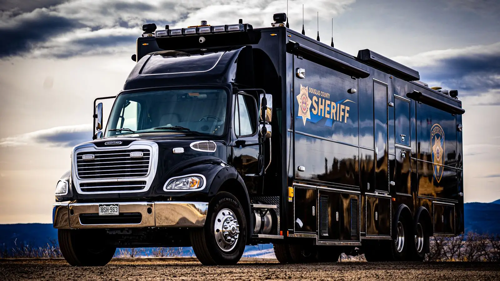 A large black sheriff’s department mobile command truck with logos, parked outdoors on a dirt surface with a cloudy sky in the background.