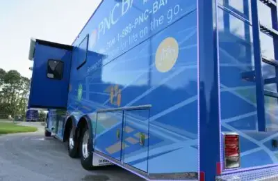 A large blue PNC Bank mobile branch vehicle is parked on a road, featuring promotional graphics, contact information, a bicycle icon, and small open compartments on the side. Trees and grass are in the background. PNC Bank