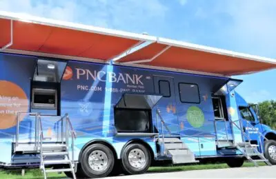 A large PNC Bank mobile branch truck with extended orange awnings is parked outdoors. The truck features stairs leading to the entrance and displays branded graphics and information on its side. PNC Bank