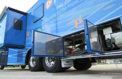 A blue banking truck labeled PNC is parked with its storage compartments open, revealing equipment and cables inside. The vehicle features shiny metal doors and branding related to mobile banking services. PNC Bank