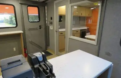 Interior of a mobile office or service vehicle with a counter, check scanner, printer, cabinets, a door, a window, and a fire extinguisher mounted near the exit.
