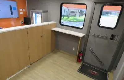 Interior of a small mobile office or vehicle with wood cabinets, a countertop, two windows, a door with metal handles, a fire extinguisher, and a floor mat that says “WATCH YOUR STEP.”.
