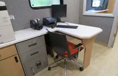 A small office workspace with a desk, black office chair, computer monitor, keyboard, printer, telephone, and a secure filing cabinet with a combination lock. The desk is near a window.