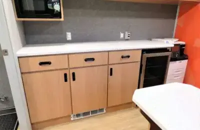 A kitchenette with light wood cabinets, a white countertop, a microwave, a small refrigerator, a printer, and electrical outlets on the wall. The floor is light wood, and one wall is orange.