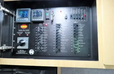 Electrical control panel with labeled switches, circuit breakers, warning labels, gauges, and indicator lights, mounted on a wooden surface with a hinged door partially open on the left side.