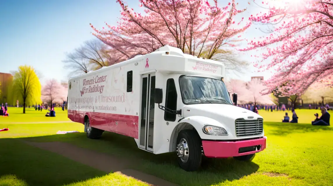 A white and pink mobile medical clinic is parked on green grass in a park with blooming cherry blossom trees under a clear sky. The side of the vehicle has text related to breast cancer screening services.
