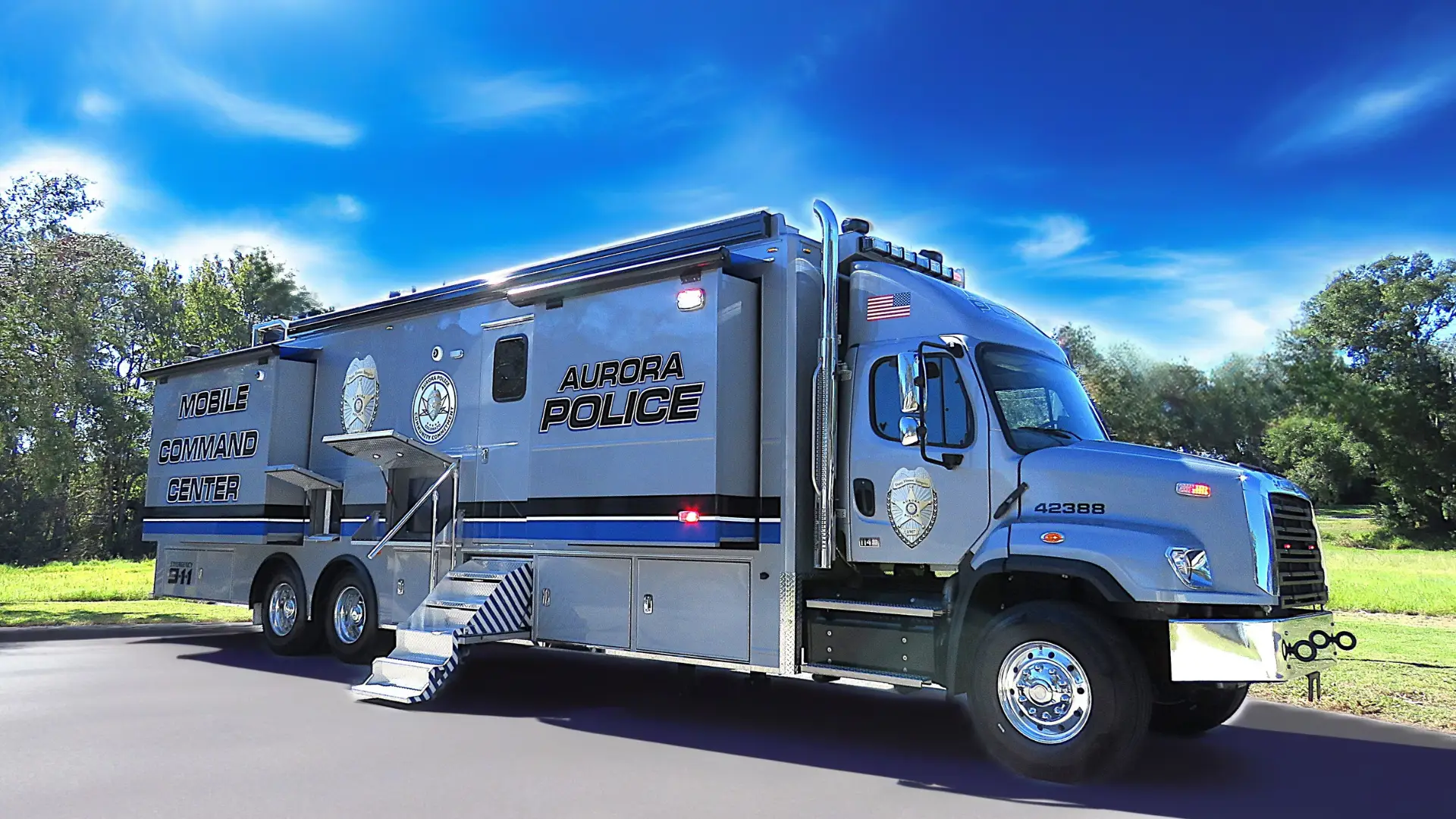 A large, silver Aurora Police mobile command center truck is parked on a paved surface near a grassy field and trees under a clear blue sky.
