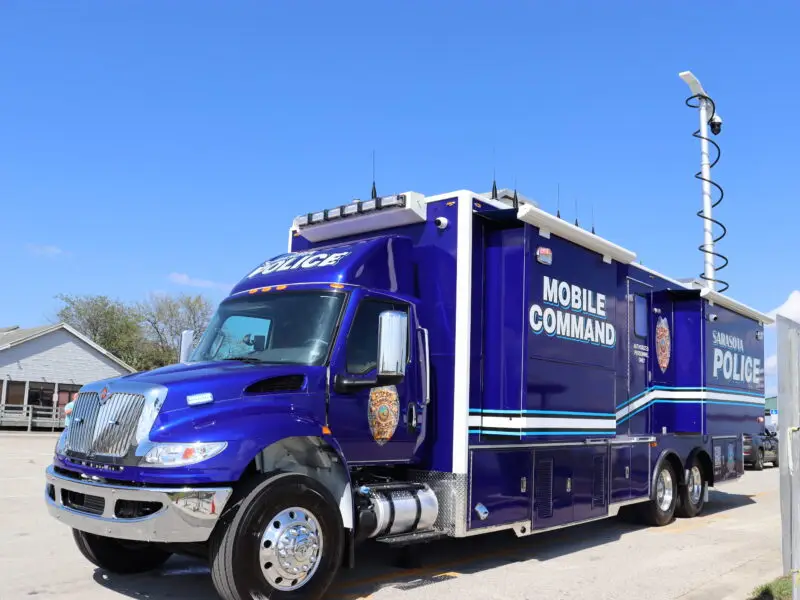 A large blue police mobile command vehicle with “Mobile Command” and “Police” written on the side is parked on a paved area. The vehicle has a raised communications mast, and buildings are visible in the background under a clear sky. Sarasota Police Mobile Command