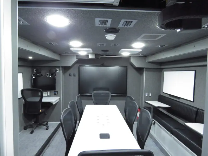 A modern conference room with a long white table, black chairs, wall-mounted monitors, whiteboards, office equipment, and built-in bench seating. The room has grey walls and ceiling lights. Sarasota Police Mobile Command