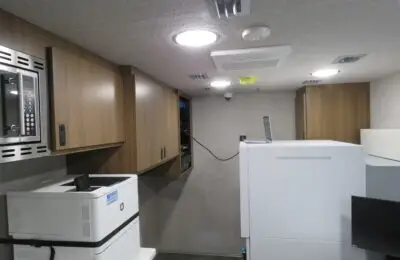A small office space with wood cabinets, a wall-mounted microwave, a printer, a copier, a laptop on top of equipment, and a monitor on a desk. The ceiling has round lights and air vents.