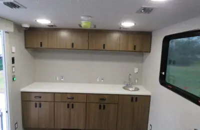 A small kitchenette with wood cabinets, white countertop, a round metal sink, and built-in overhead cabinets. The space is well-lit with ceiling lights and has a large window on the right side.