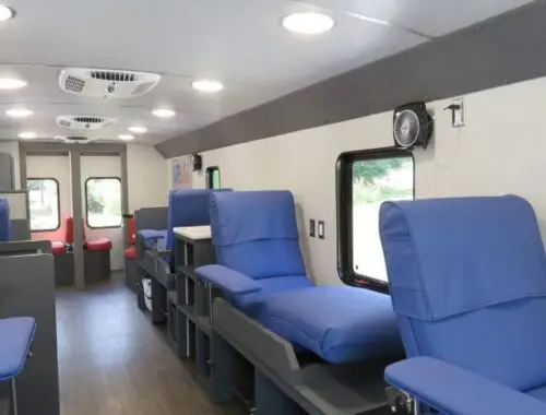 Interior of a mobile medical unit with several blue reclining chairs arranged along the wall, small tables beside each chair, white ceiling lights, and a wood-look floor. Windows line one side of the vehicle. OneBlood