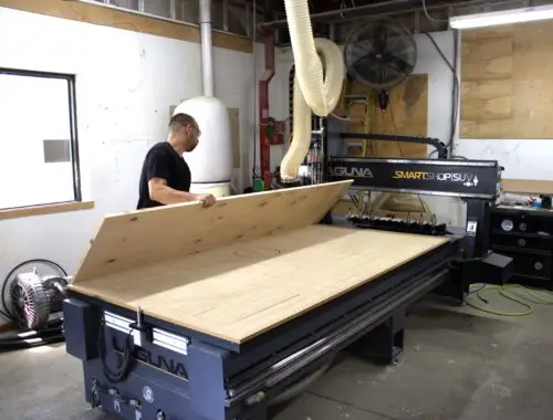 A man places a large sheet of plywood onto a CNC router table in an MBF Industries company workshop, preparing the material for cutting. Various workshop equipment and tools are visible in the background.