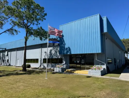 A blue and gray industrial building with large windows and a flat roof, a U.S. flag on a flagpole in front, and a grassy lawn with a tree on a clear, sunny day.