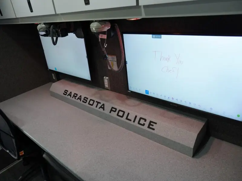 A workstation with two large monitors, a chair, and a countertop labeled Sarasota Police. One monitor displays a handwritten message that says, Thank you Chief! The surroundings appear organized and modern.