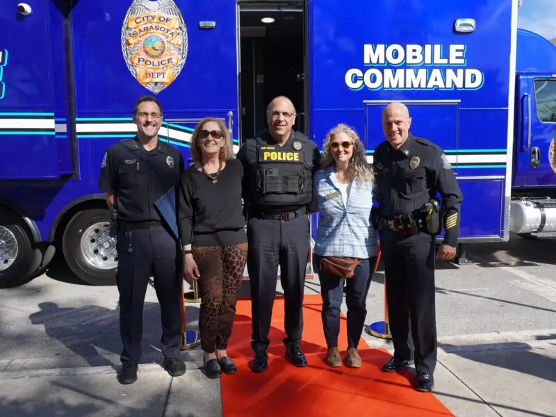 Five people stand and smile on a red carpet in front of a blue “Mobile Command” police vehicle. Three are in police uniforms, and two are in casual clothes. The “City of Sarasota Police Dept” badge logo is visible on the vehicle.