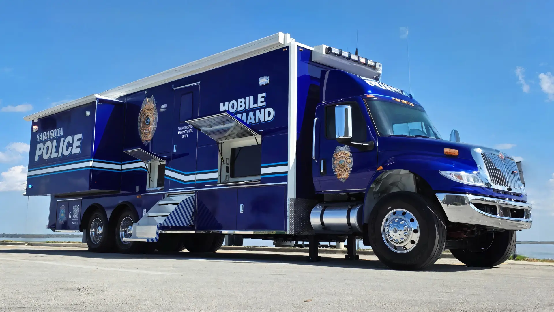 A large blue Sarasota Police mobile command vehicle is parked on pavement under a clear sky. The truck features department logos, labeled compartments, an open door, and extended stairs.
