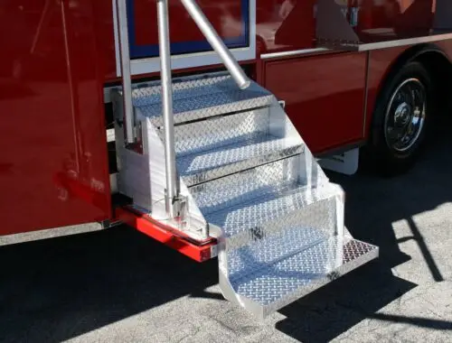 A set of shiny, metal diamond plate steps with a handrail is attached to the side of a red vehicle, positioned on a paved surface. OneBlood