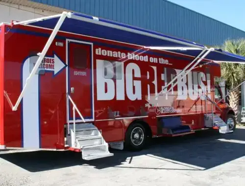 A large red mobile blood donation bus with the words “donate blood on the BIG RED BUS” parked outside a building, with awnings extended and a ramp leading to the entrance. OneBlood