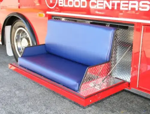 A blue cushioned bench seat extends from the side of a red blood donation vehicle, with metal diamond plate panels surrounding the seat.