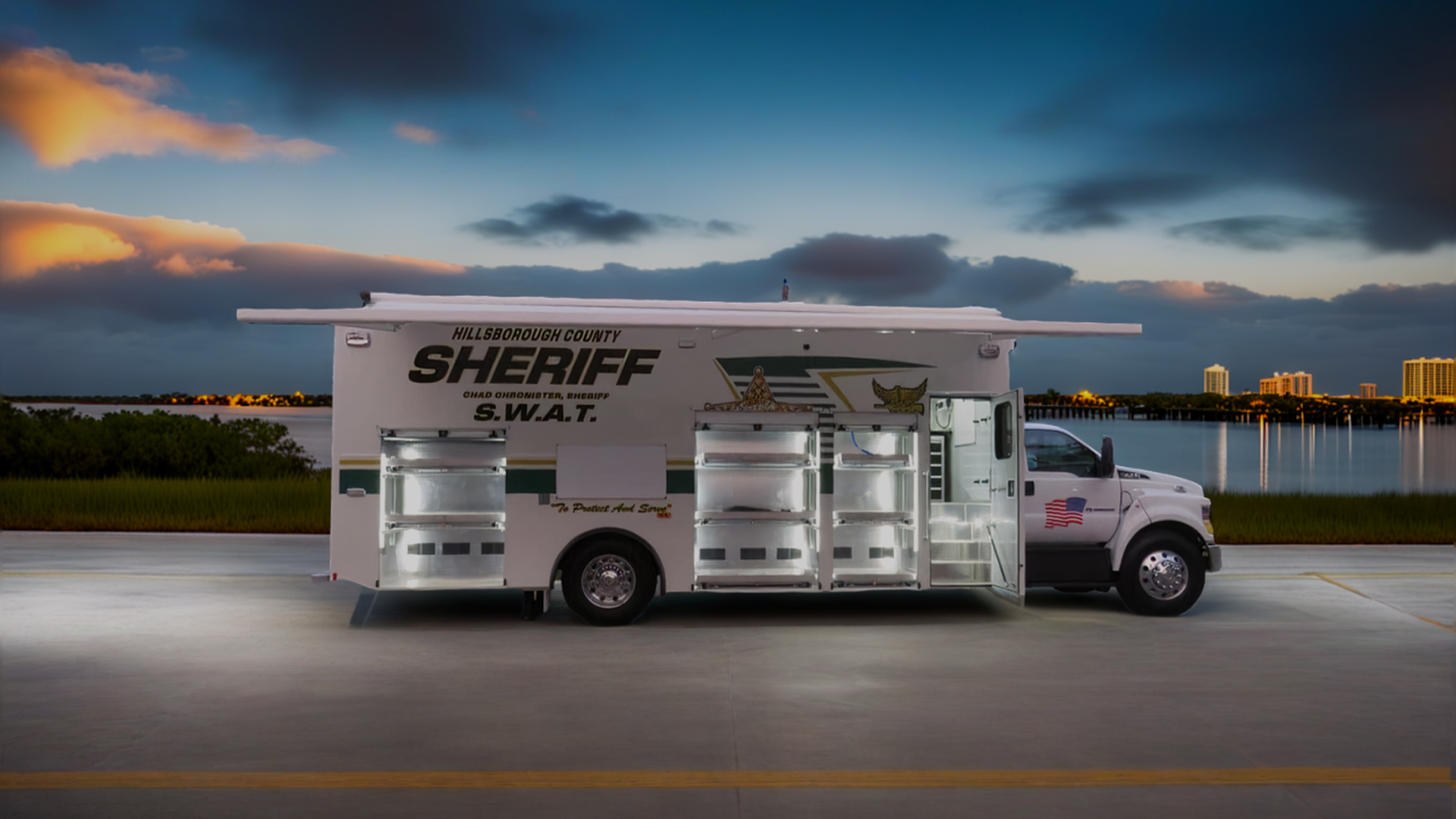 A Hillsborough County Sheriff S.W.A.T. vehicle with multiple open compartments is parked by a waterfront at dusk, with city buildings visible in the background.