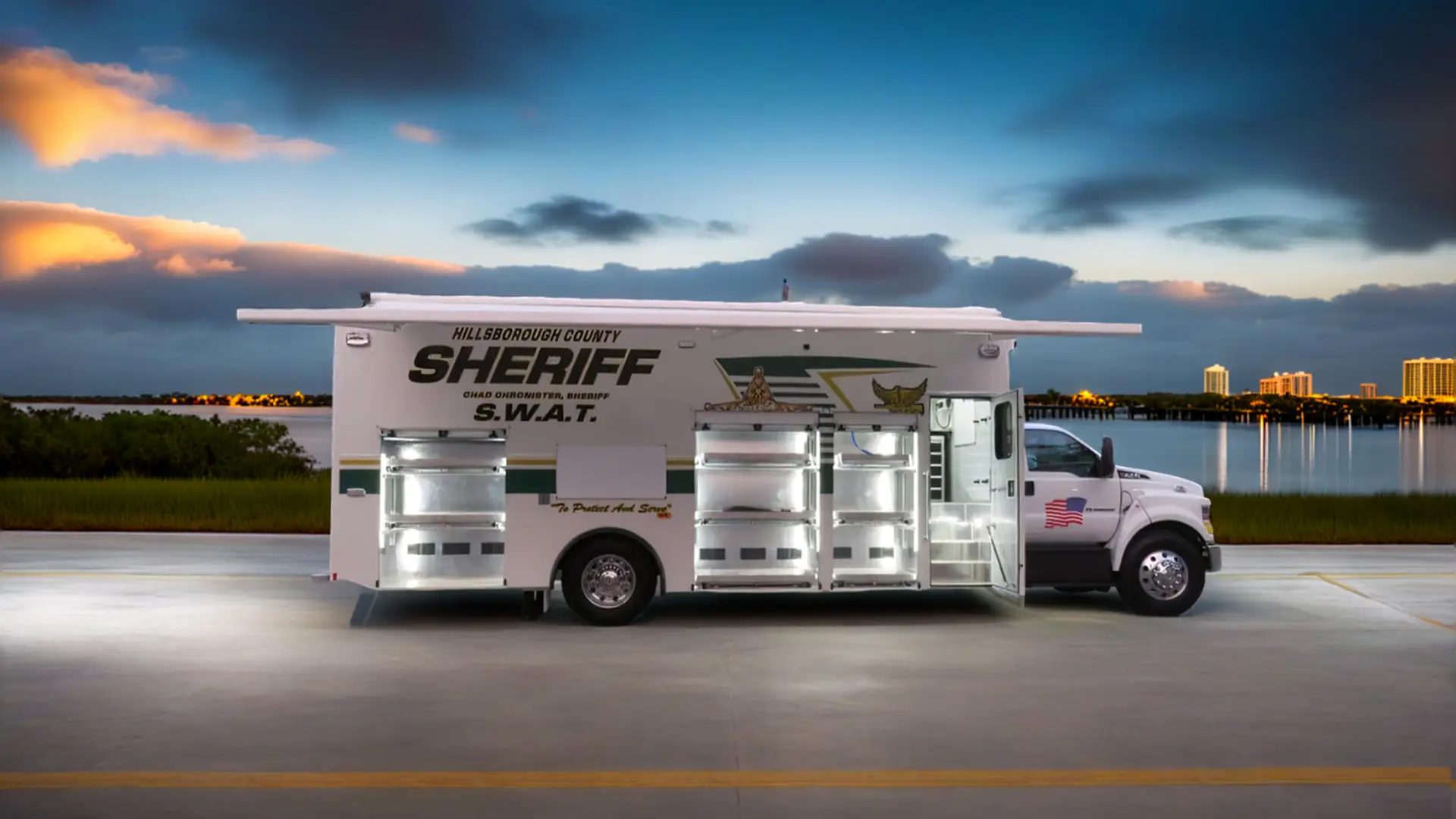 A large Hillsborough County Sheriff S.W.A.T. vehicle is parked on a roadway near a body of water at dusk, with several side compartments open and city buildings visible in the background.