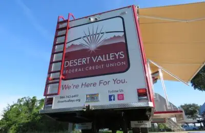 A mobile unit for Desert Valleys Federal Credit Union is parked outdoors. The back features the credit union’s logo, contact information, stairs, and a red ladder. An awning extends from the side.