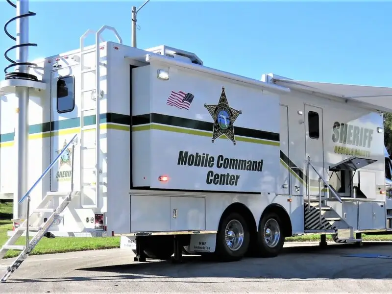 A Volusia County Sheriffs Office Public Safety Vehicle serves as a mobile command center, parked on a road with its side awning extended, stairs to the entrance, a tall mast raised from the roof, and prominent law enforcement insignia.