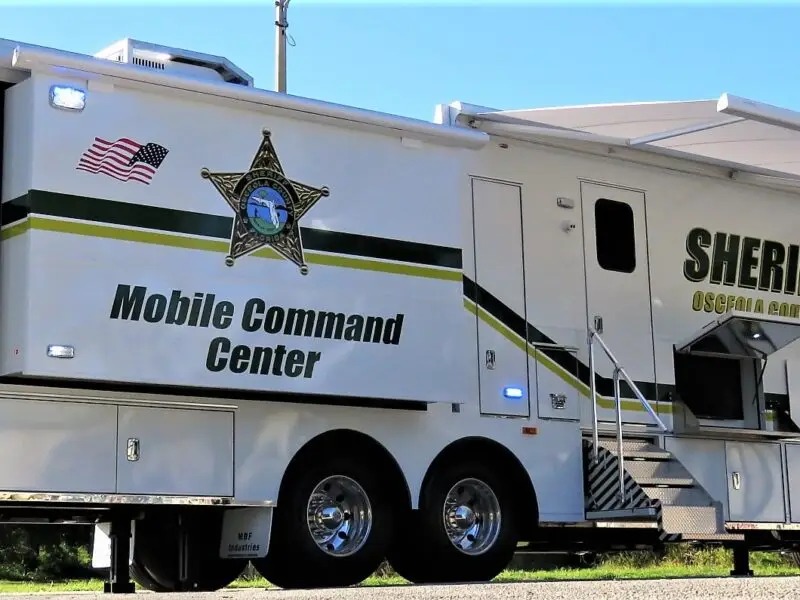 A large white Volusia County Sheriffs Office Public Safety Vehicle mobile command center is parked outdoors, featuring official markings, an extended awning, side stairs, and attached equipment. Trees and grass are visible in the background.
