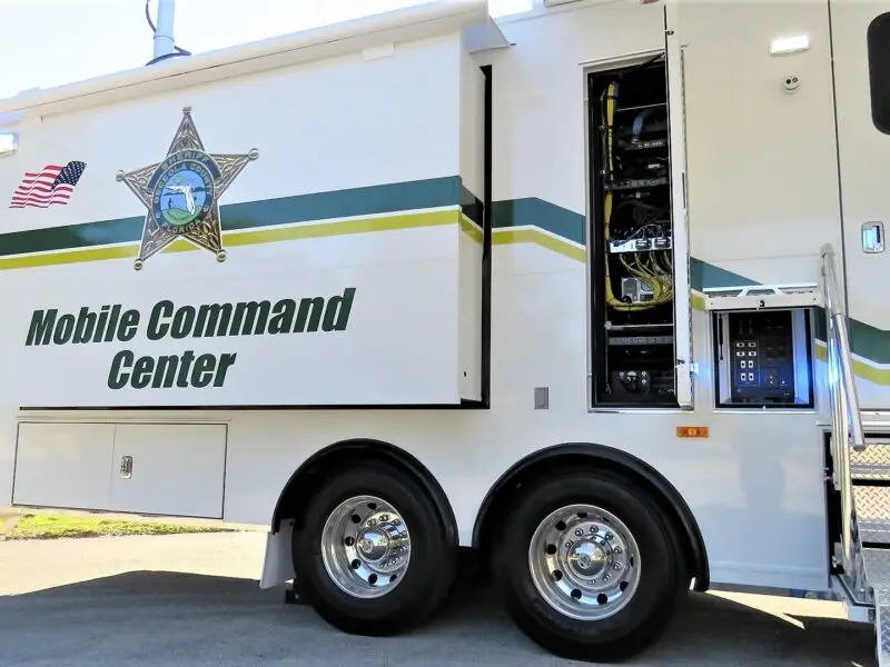 A large white Volusia County Sheriffs Office Public Safety Vehicle labeled Mobile Command Center with official insignia, an American flag, and electronic equipment visible inside. Metal stairs lead up to an entrance on the right side.
