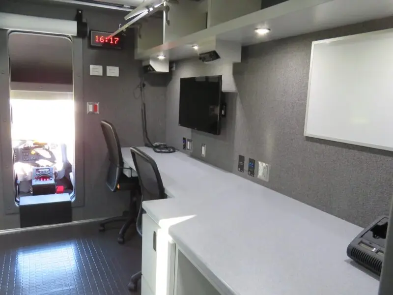A modern Volusia County Sheriffs Office Public Safety Vehicle interior featuring a long counter with chairs, a mounted monitor, whiteboard, wall outlets, and equipment. A digital clock shows 16:17, with the driver’s cab visible in the background.