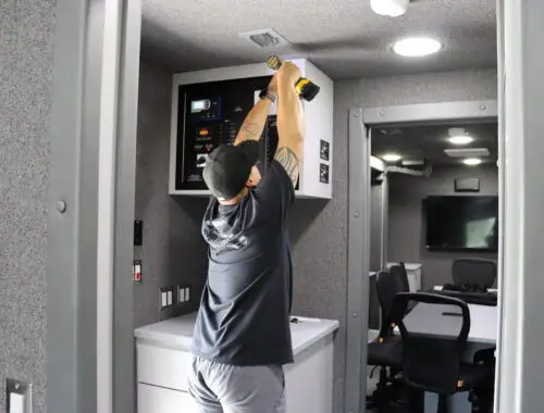 A person wearing a black shirt and cap uses a power drill to work on a panel above a countertop in a gray, modern MBF Industries company office or control room. Office chairs and a monitor are visible in the background.