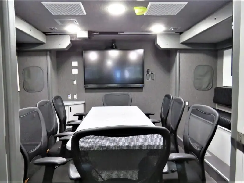A conference room with a long white table, eight black mesh chairs, a large wall-mounted screen, control panels, and built-in cabinetry in a gray color scheme—styled for the Volusia County Sheriffs Office Public Safety Vehicle team.