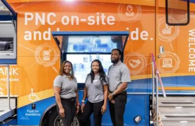 Three people wearing gray shirts stand in front of a brightly colored PNC Bank mobile branch with an open window, informational screens, and visible accessibility signage. PNC Bank
