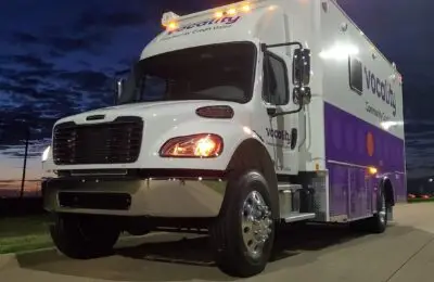 A white and purple mobile service truck with vocality Community Credit Union written on the side is parked on a paved surface at dusk, with headlights on and a partly cloudy sky in the background.