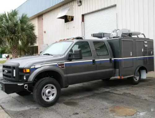 A large gray utility truck with a crew cab and extended cargo compartments is parked outside a beige industrial building with a roll-up door. A palm tree is visible in the background.
