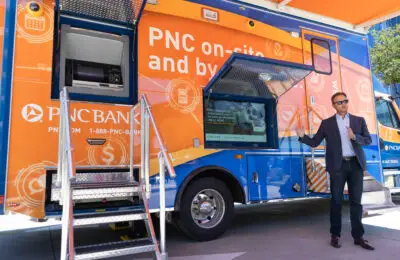 A man in a suit stands beside a brightly colored PNC Bank mobile branch truck with open service windows, stairs leading up, and signage displaying “PNC on-site and by appointment.”. PNC Bank