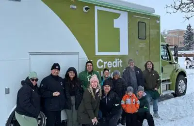 A group of people in winter clothing pose and smile in front of a green Credit 1 truck on a snowy day, with snow covering the ground and trees in the background.